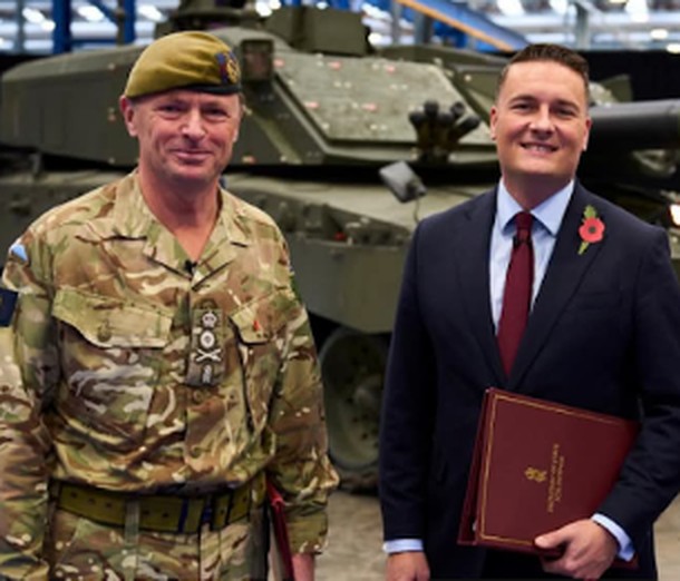 A British Army officer in camouflage uniform stands beside a government minister holding a red folder, in front of a tank.